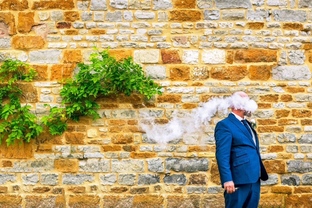 A man in a bright blue suit stands against a yellow stone wall, holding a vape in one hand. A thick cloud of white vapor covers his face and trails sideways across the frame. A small patch of green leaves grows from the wall beside him.