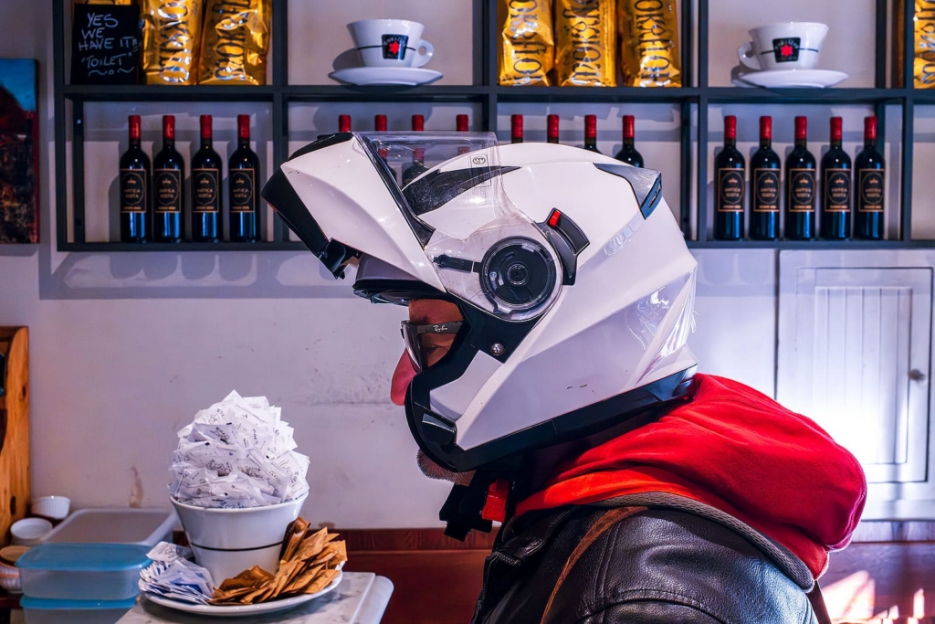 A person sits indoors wearing a large white motorcycle helmet, a red hoodie, and a black leather jacket. Behind them, shelves are lined with wine bottles, gold coffee bags, and oversized coffee cup decorations, creating a strange mix of café and biker imagery.