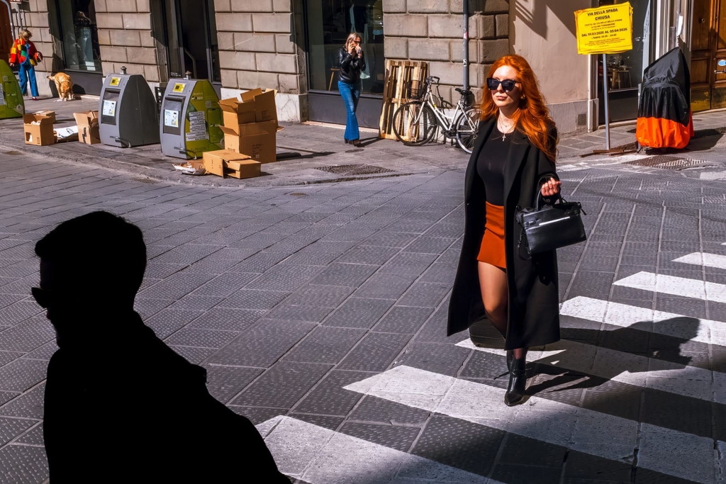 A woman with long red hair walks confidently across a crosswalk in bright sunlight, wearing a black coat, sunglasses, and an orange skirt. In the foreground, a dark silhouette of a person partially blocks the view, creating strong contrast against the lit street scene.