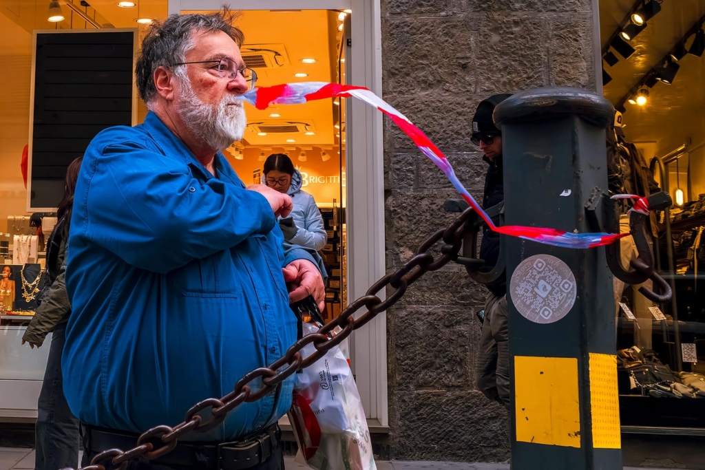 An older man in a blue shirt stands on a street holding a plastic bag while pulling a red-and-white tape tied between posts. The tape stretches across the frame, creating a diagonal line in front of him. Behind him, people stand near a brightly lit shop entrance.