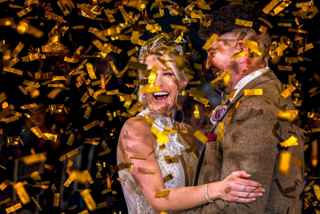A bride and groom embrace closely, both smiling and laughing as golden confetti rains down around them.
The bride’s white dress and sparkling headpiece contrast with the groom’s textured suit, set against a dark background that makes the falling confetti glow.