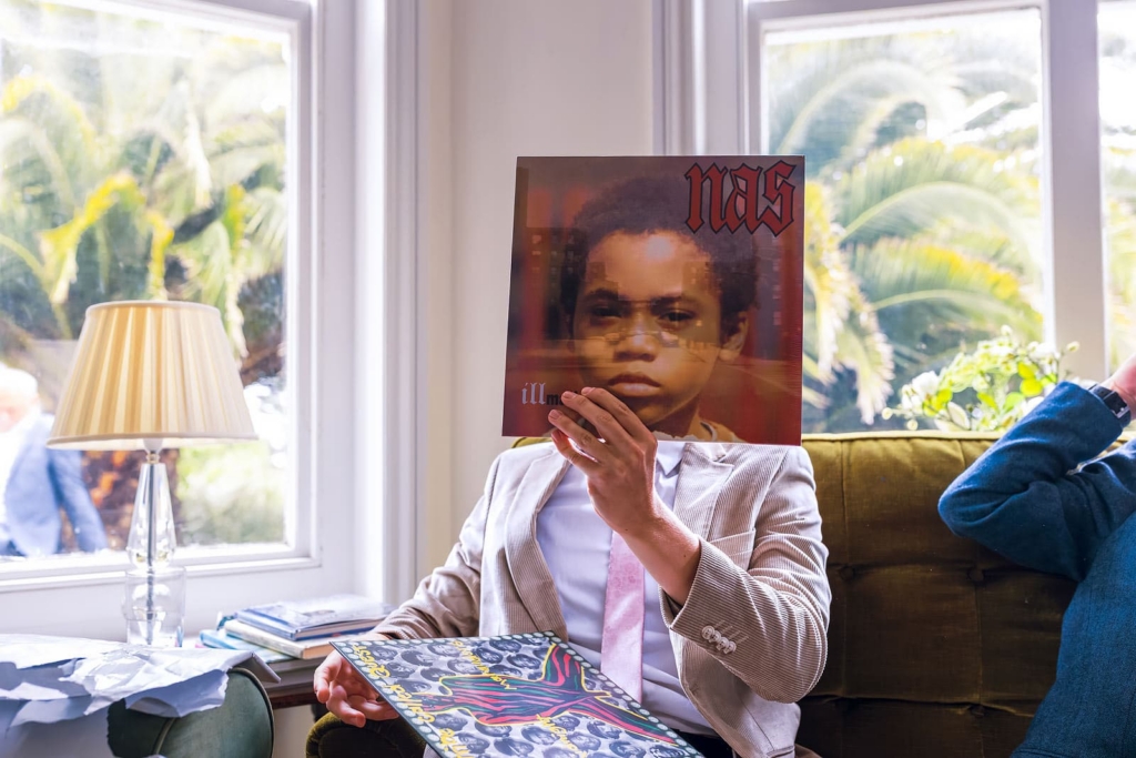 A young child in a pale suit and pink tie sits in an armchair indoors, holding a vinyl record sleeve in front of their face so the album portrait lines up with their body. Sunlight pours through tall windows behind them, while another record rests across their lap.