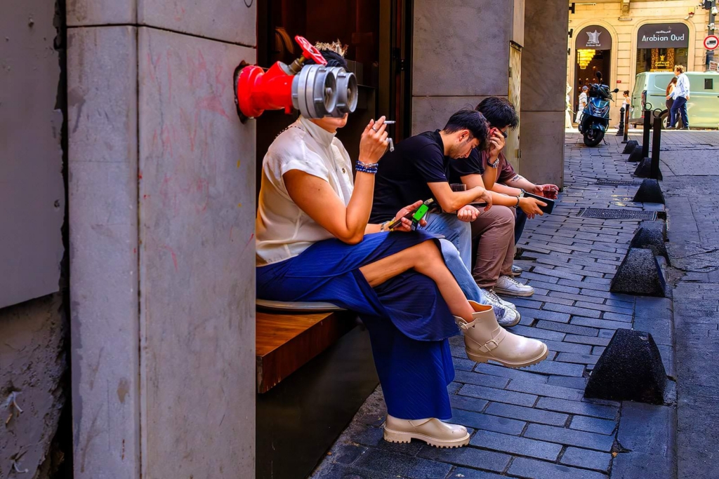 A person sits on a narrow city sidewalk bench with their head aligned perfectly behind a red wall-mounted pipe, making it look like a mechanical fixture replaces their face. Two others sit beside them, looking down at their phones, while a scooter and pedestrians appear further down the street.