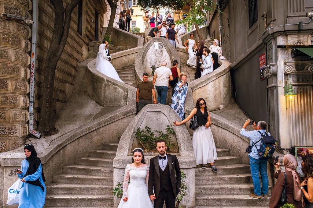 A bride and groom stand hand in hand at the bottom of a large, sculptural stone staircase in a busy city passage. Around them, pedestrians, tourists, and other couples move through the steps, creating a striking contrast between the formal wedding portrait and the everyday urban crowd.