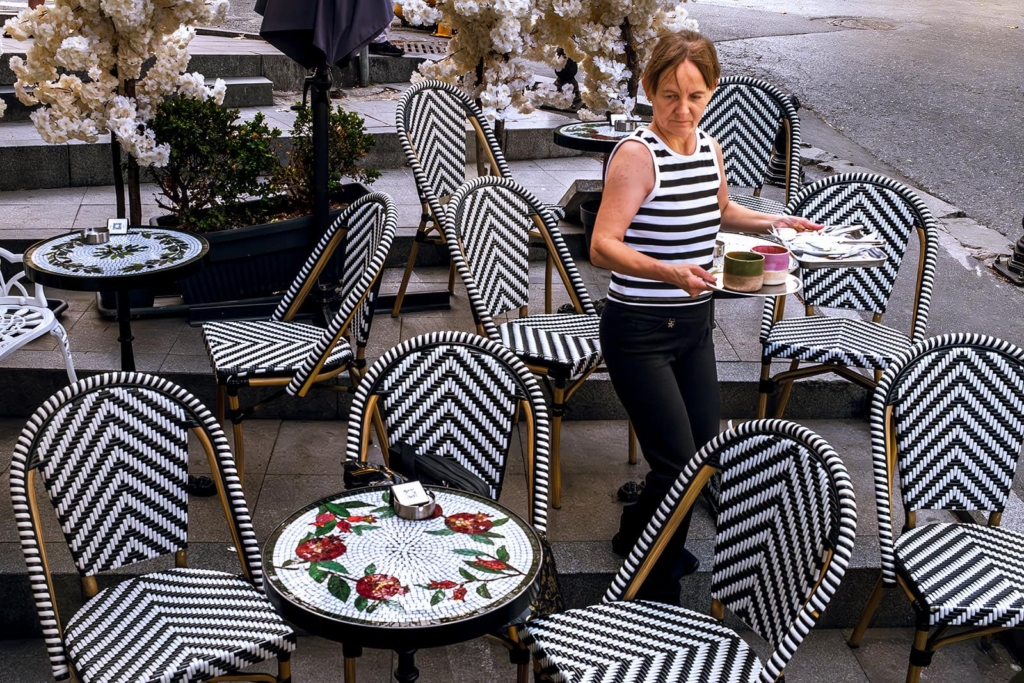 A woman in a striped sleeveless top walks through an outdoor café carrying trays with cups and plates. Around her, black-and-white patterned chairs and small mosaic tables create a busy geometric scene against pale flowers and stone steps in the background.