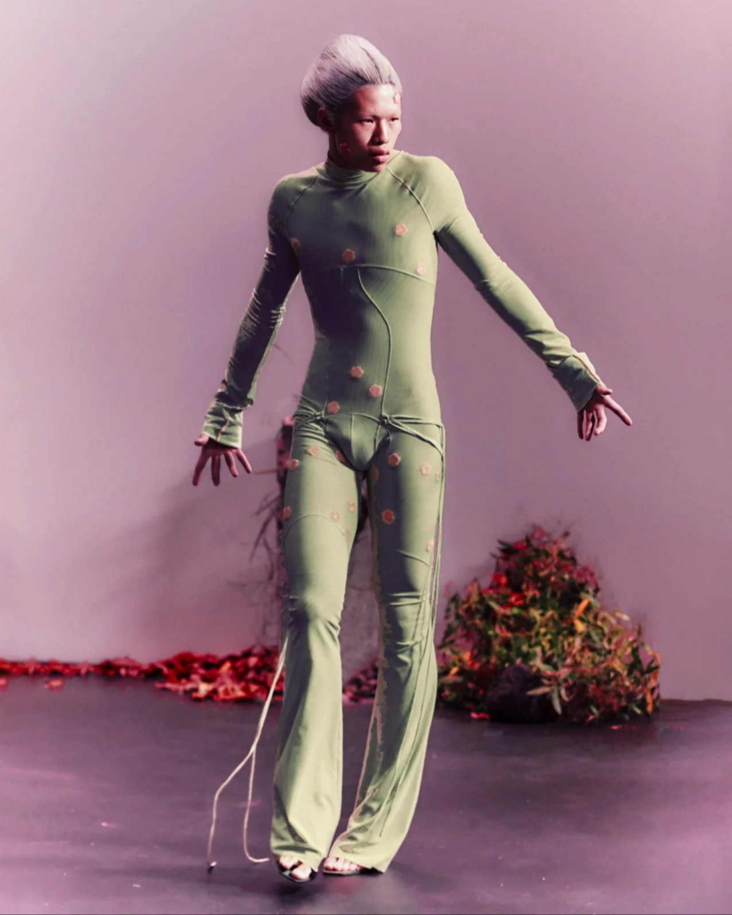 A runway image of a model in a fitted pale green bodysuit, posed mid-step against a soft pink-lit background. The look feels futuristic and slightly surreal, with the body held in a tense, almost sculptural pose.