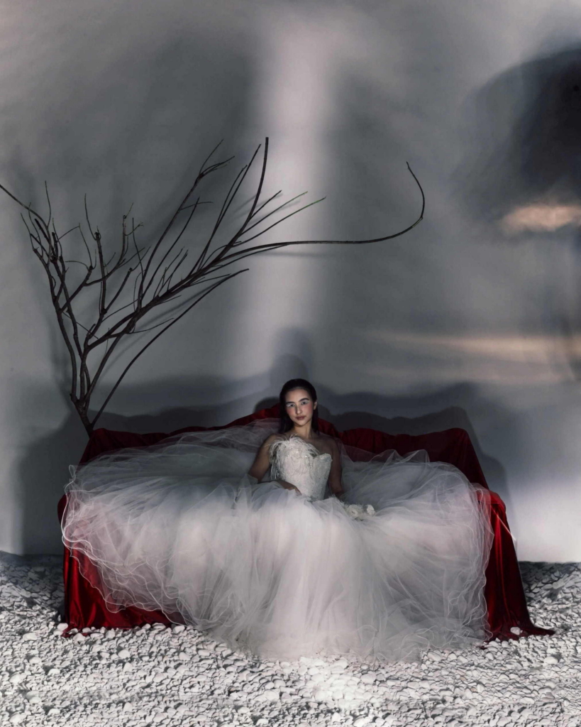 A woman in a voluminous white gown sits on a deep red sofa in a dramatic studio set. Bare branches, white stones, and projected shadows create a surreal, theatrical atmosphere around her.