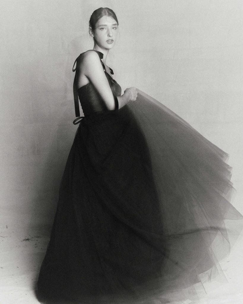 A timeless black-and-white fashion portrait that leans into restraint and quiet elegance. The soft lighting and subtle grain give the image a film-like quality, while the voluminous sheer skirt introduces movement and depth. The pose feels poised yet unforced, allowing the texture of the fabric and the simplicity of the composition to carry the image.