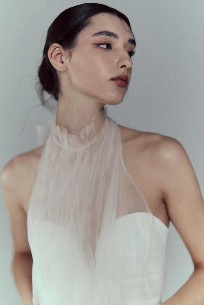 A soft portrait of a woman in a white dress with a sheer high neckline, photographed against a pale background. Her face is turned slightly to the side, giving the image a quiet, elegant stillness.
