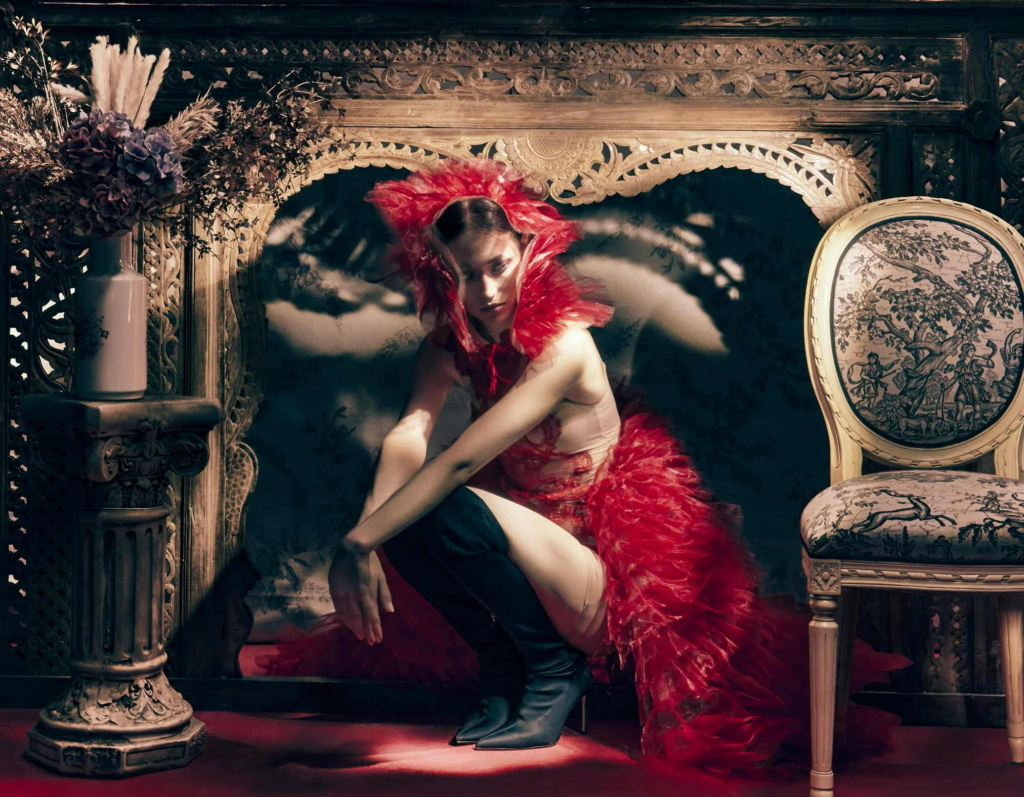 A fashion portrait of a model in a dramatic red feathered garment, crouched within an ornate, antique interior. The scene feels theatrical and painterly, with rich textures and controlled light.
