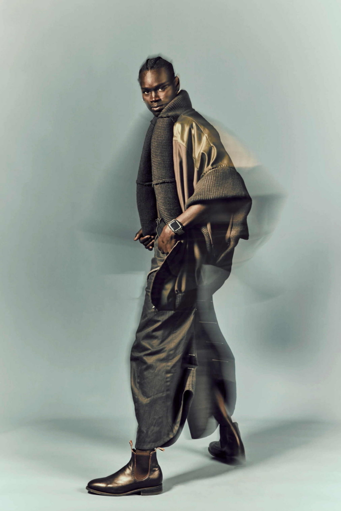A fashion portrait of a model in layered brown styling, captured mid-step against a pale studio background. A slight motion blur adds movement and softness to the otherwise controlled image.