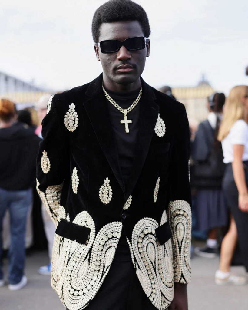 A street-style portrait of a man in dark sunglasses and an embroidered black velvet blazer. The image feels polished and self-possessed, with the crowd behind him softly out of focus.