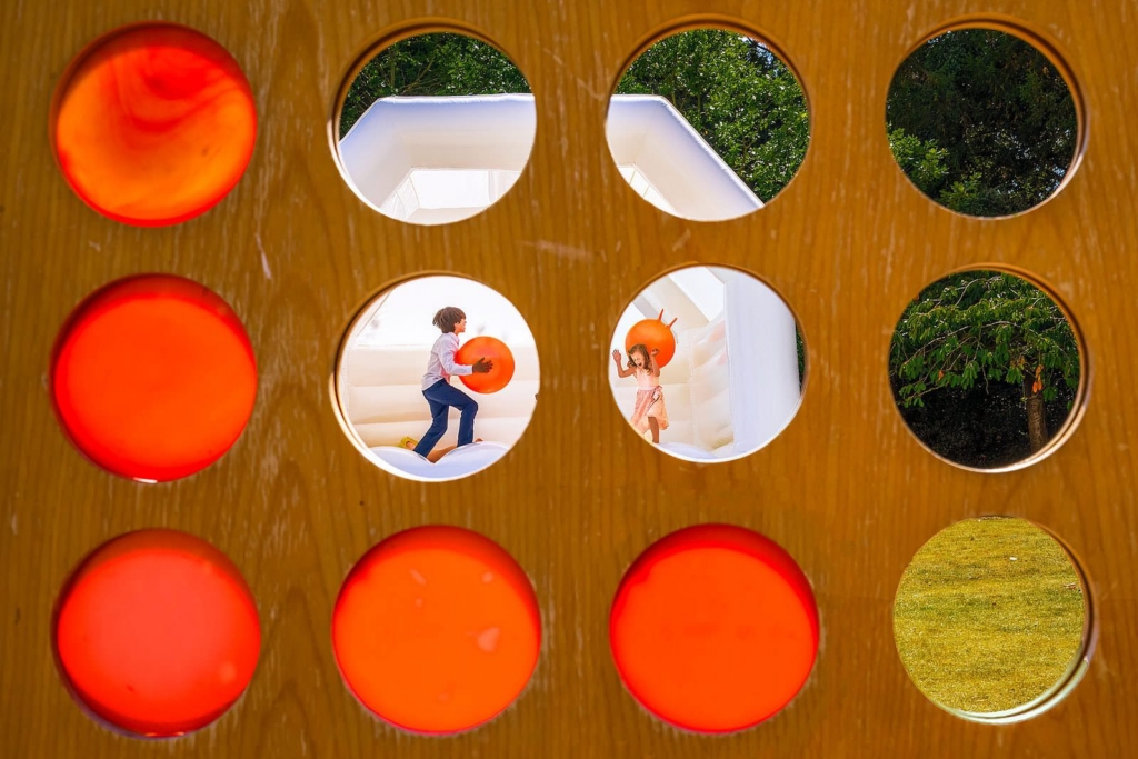 Two children are seen through circular holes in a wooden game board, playing inside a white inflatable structure. Several of the holes are filled with bright orange balls.