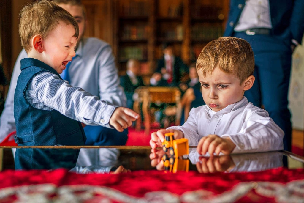 Two young boys stand at a reflective table indoors, focused on a small yellow toy truck between them. One points excitedly while the other looks down with a serious, concentrated expression. The warm room behind them is softly blurred, with adults and shelves in the background.
