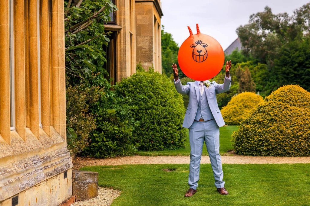 A man in a light blue suit stands on a lawn outside a stone building, holding a large orange balloon that completely covers his face. His arms are raised as if catching or tossing it.