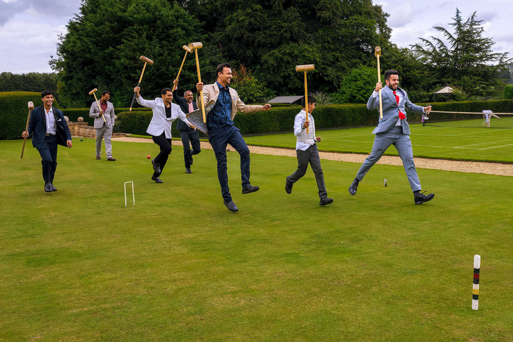 A group of men in suits run across a manicured lawn holding croquet mallets in the air, laughing and mid-stride as if charging into a game. The formal clothing and playful movement create a lively, celebratory scene in an elegant garden setting.