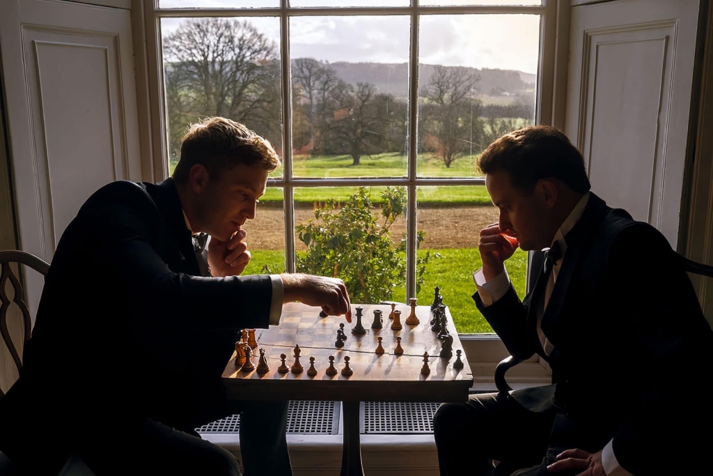 Two men in black formalwear sit across from each other at a small table, deeply focused on a game of chess. They are silhouetted against a large window overlooking a bright green landscape, giving the quiet scene a dramatic, thoughtful mood.