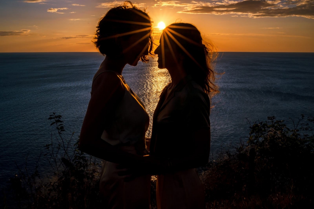 Two women stand close together in silhouette at the edge of the sea during sunset, their faces nearly touching. The low sun sits between them, sending bright rays across the sky and reflecting a golden path over the water behind them.