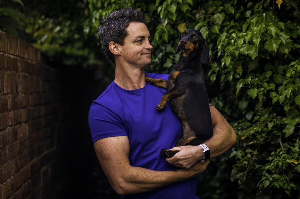 Dan Morris, the photographer, stands outdoors holding a black-and-tan dachshund in his arms. He wears a bright blue T-shirt and looks at the dog while the dog looks slightly off to the side. They are framed by a brick wall on one side and dense green leaves on the other.