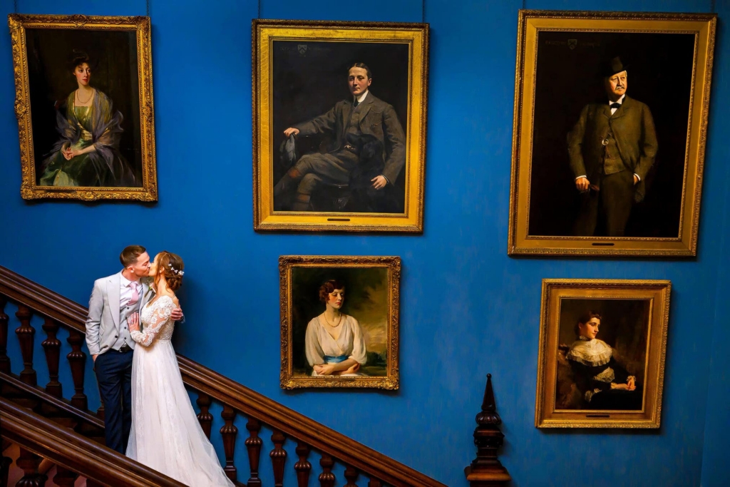 A bride and groom kiss on a dark wooden staircase beneath a wall of large framed portraits. Their small figures sit in the lower corner of the scene, surrounded by a striking blue wall and formal painted faces watching from above.