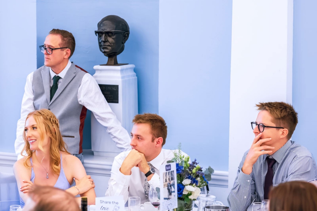 A group of wedding guests sit around a table indoors, dressed in formal attire, all looking toward something happening off-frame. Behind them, a dark bust sculpture on a pedestal mirrors their gaze, adding a quiet, slightly humorous echo to the scene.