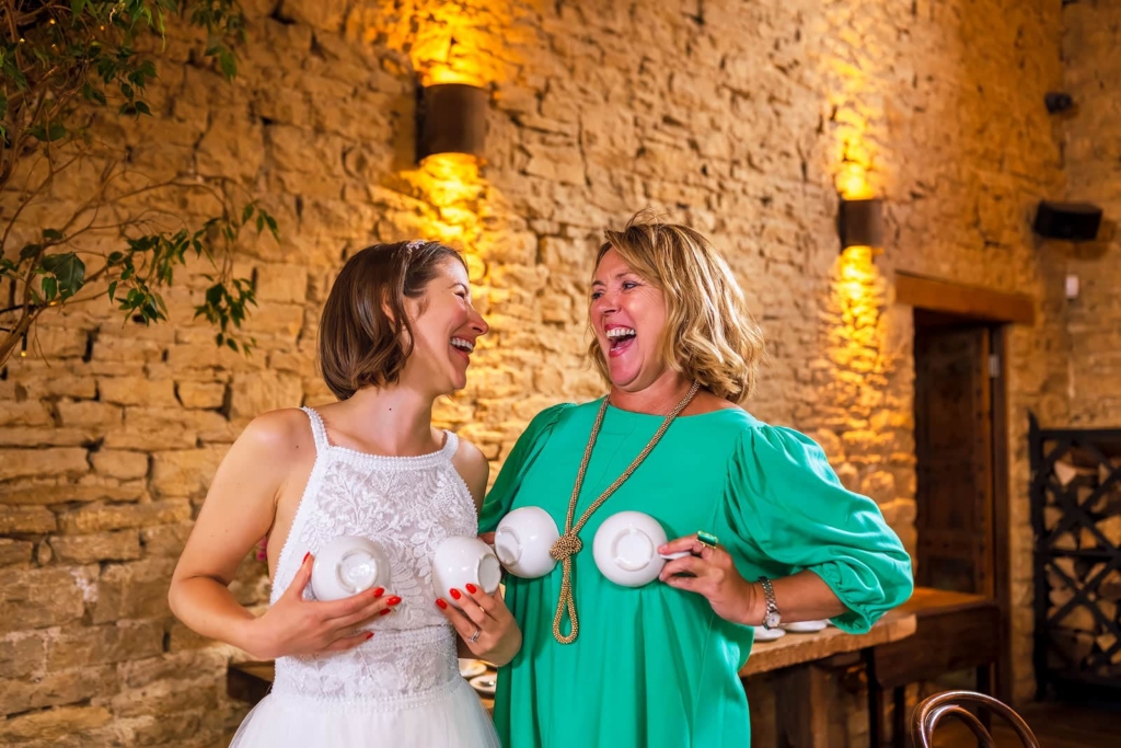 Two women stand laughing together indoors against a warm stone wall, each playfully holding white teacups across their chest like a joke prop. The image captures a candid, mischievous moment of shared humor during what appears to be a wedding celebration.