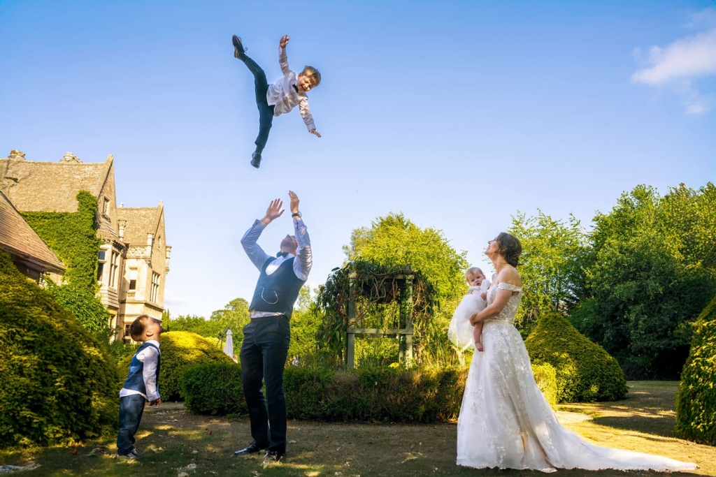 A man tosses a young boy high into the air in a garden, while another child watches from below. A bride holding a baby looks on, smiling, as the boy reaches the peak of the throw against a clear blue sky. A large house and green hedges frame the scene in warm daylight.