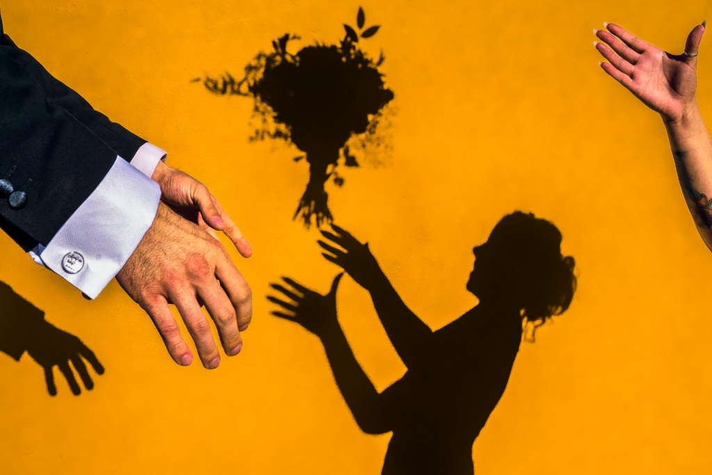 A sharp shadow of a bride appears on a bright yellow wall, her hands raised as if tossing a bouquet into the air. In the foreground, only parts of two real people are visible—one man’s hand in a dark suit sleeve and another raised hand reaching in from the right.