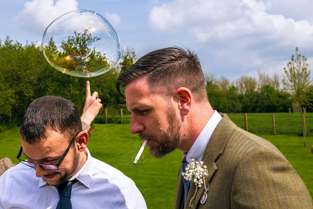 Two men stand closely together outdoors on a bright green lawn, while a large soap bubble floats above them. One man looks down, and the other, wearing a boutonniere and a cigarette in his mouth, faces sideways. In the background, a hand reaches up with one finger pointing toward the bubble.