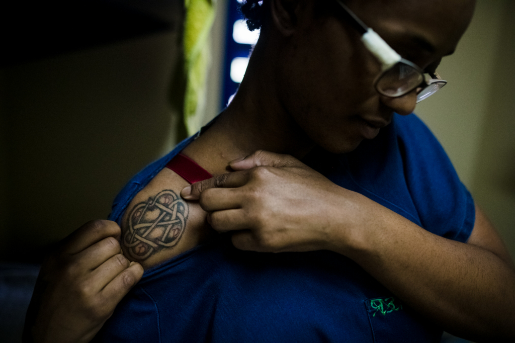 Close-up documentary photograph of a woman inside a Barbados prison revealing a tattoo beneath her uniform shirt.