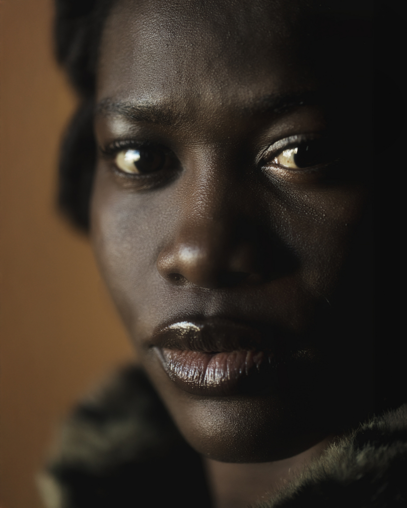 A close-up portrait of a person with deep brown skin, softly illuminated by warm directional light that highlights their eyes and facial features. Their steady gaze and neutral expression create an intimate, contemplative mood, while the dark background keeps the focus entirely on the face.