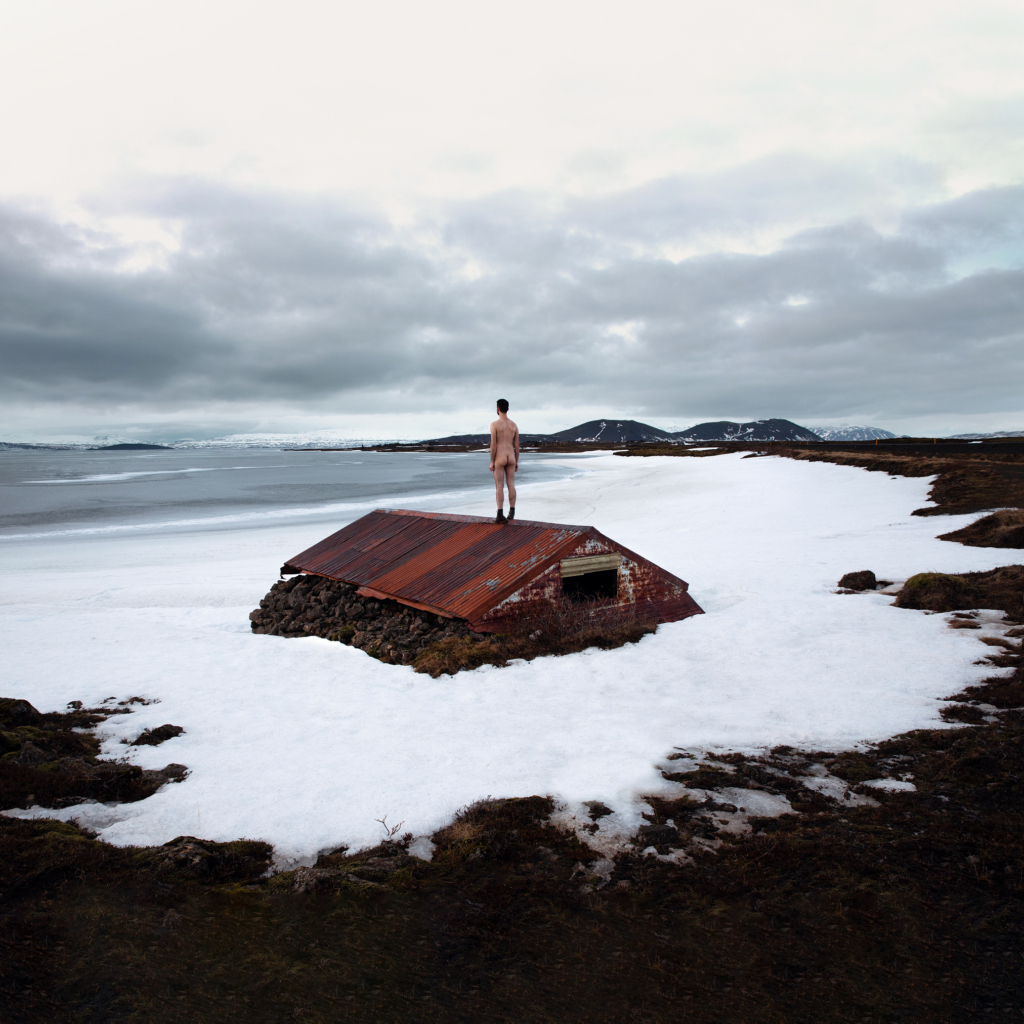 A lone figure stands on the rusted roof of a partially buried structure surrounded by snow, facing a wide coastal landscape and distant mountains. The overcast sky and expansive surroundings create a sense of isolation and stillness, while the contrast between the figure and the winter landscape draws the eye across the scene.