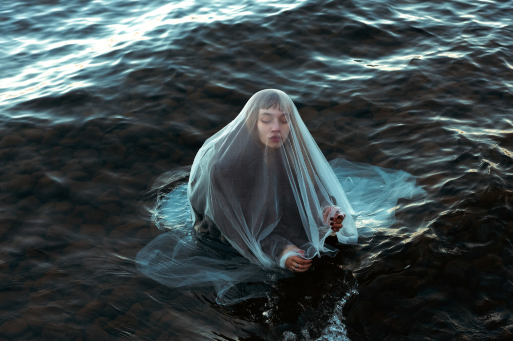 A woman sits in shallow flowing water, draped in a translucent veil that softens her features and spreads around her in the current. With her eyes closed and hands resting gently in the water, she appears calm and introspective, while the dark river and rippling surface create a quiet, contemplative atmosphere.