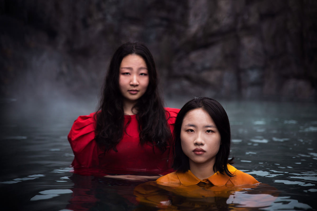Two young women stand in still water beneath mist-covered cliffs, wearing vivid red and orange garments that contrast with the dark surroundings. They look directly at the camera with calm, serious expressions, creating a sense of quiet presence and connection within the landscape.