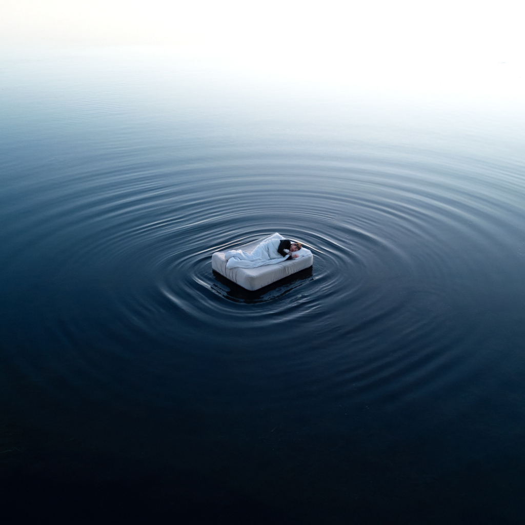 A person lies asleep on a floating mattress in the middle of a calm body of water, wrapped in white bedding. Gentle ripples spread outward in concentric circles around the bed, creating a quiet sense of stillness and isolation within the vast, open scene.