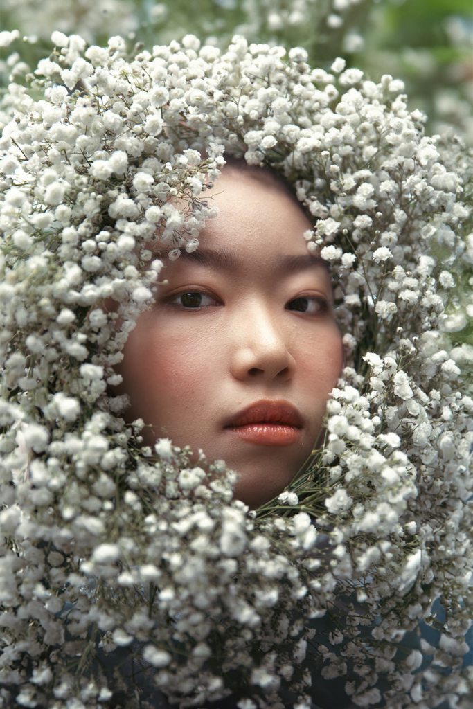AI-generated portrait of a young woman whose face is framed by a dense arrangement of small white flowers. She looks directly at the viewer with a calm, neutral expression, while the soft natural light and blurred greenery create a delicate and serene atmosphere.