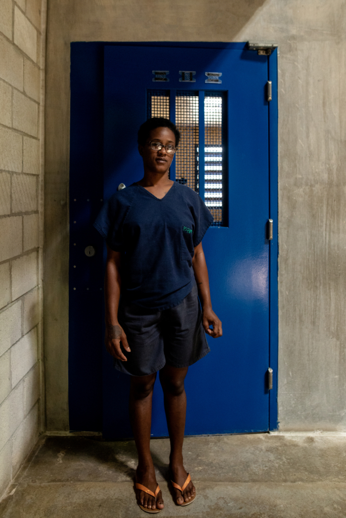 Portrait of a woman standing barefoot in front of a blue prison cell door inside a Barbados correctional facility.