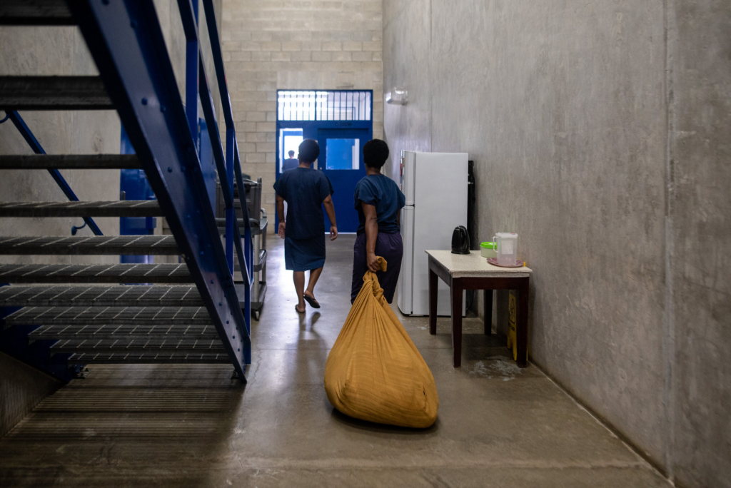 Two people in blue uniforms walk through a stark concrete hallway inside a prison facility, while one drags a large yellow laundry bag across the floor. Harsh overhead light, metal stairs, and sparse furnishings emphasize the institutional setting and the quiet routine of the scene.