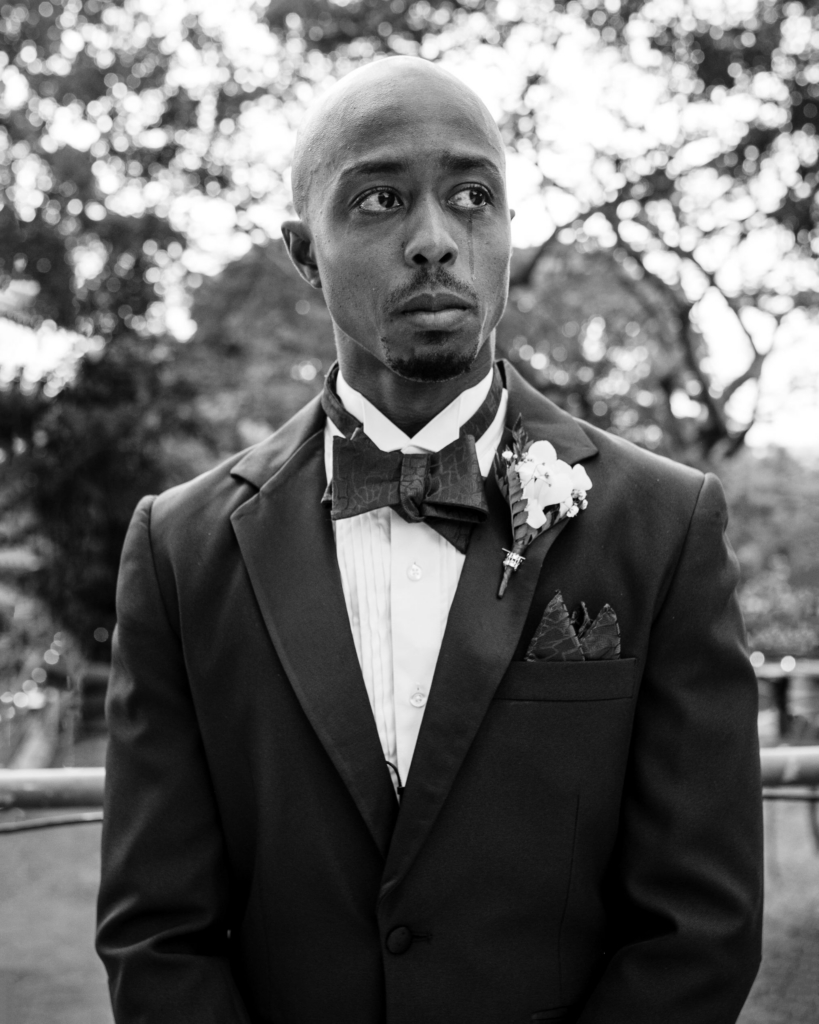 In a black-and-white portrait, a groom in a dark tuxedo stands outdoors with tears visible on his face as he looks off to the side. Blurred trees and bright light in the background contrast with his still posture and emotional expression.