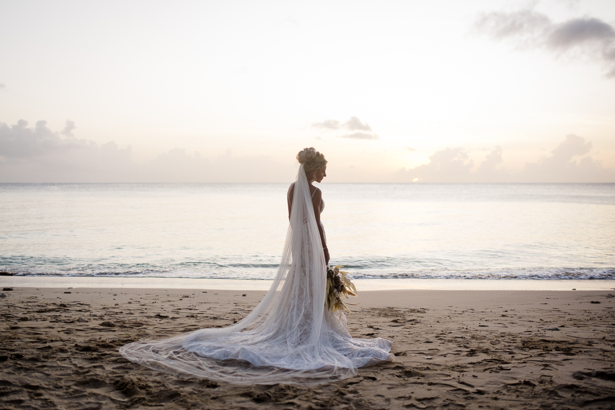 A bride stands alone on a quiet beach at sunrise, wearing a long veil that trails across the sand behind her while she holds a bouquet at her side. Soft pastel light reflects off the calm water, and her still posture facing the horizon gives the scene a peaceful, contemplative mood.