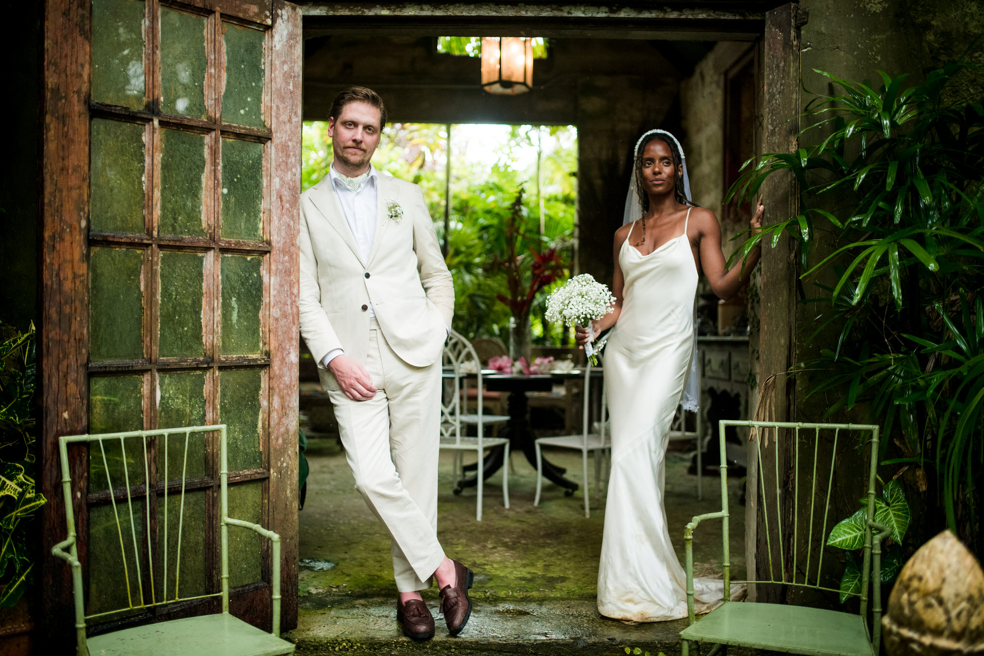 Bride and groom standing in the open doorway of a weathered tropical villa surrounded by lush green plants during an intimate wedding portrait session.