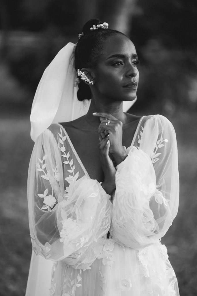 A bride in an embroidered wedding dress stands in a black-and-white portrait with her hands gently folded near her face. Her veil and pearl hair accessories frame her expression as soft light highlights the texture of the fabric and her calm, steady posture.