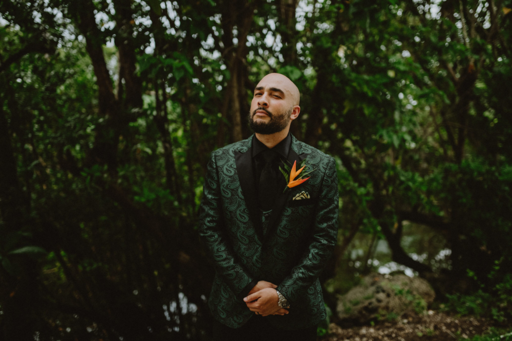 A man stands in a dense tropical forest wearing a dark patterned suit with an orange flower pinned to his jacket. The deep green foliage surrounds him closely, and his upright posture and direct expression give the portrait a calm, confident mood.