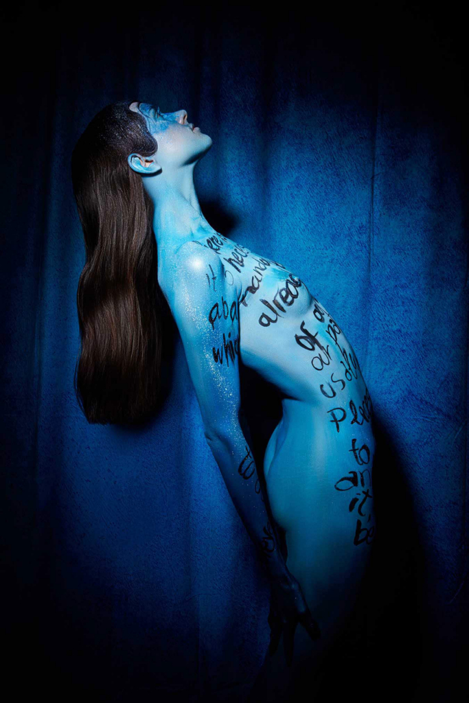 Conceptual nude portrait with handwritten typography, blue body paint, and theatrical low-key lighting.