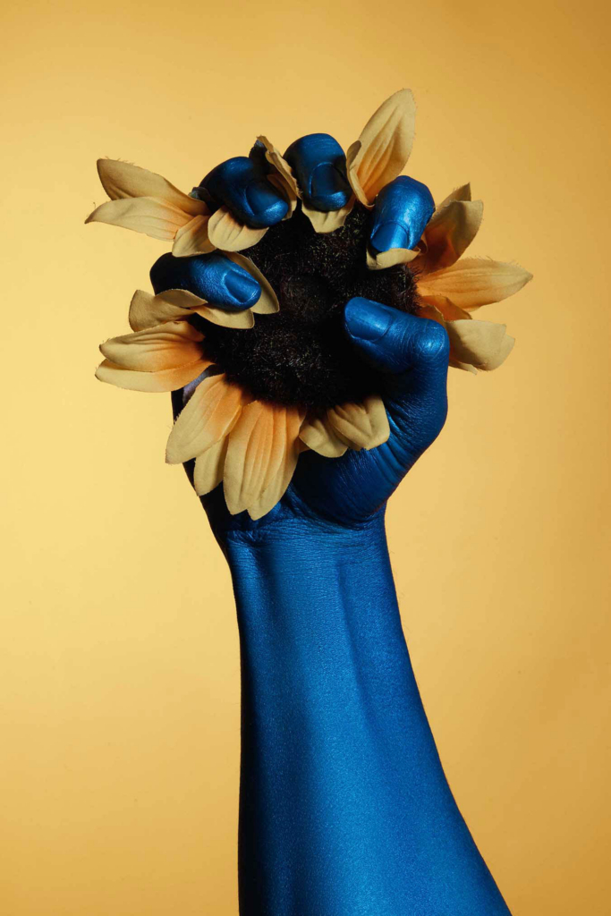 Surreal floral still life with painted blue hand and bold complementary color contrast.