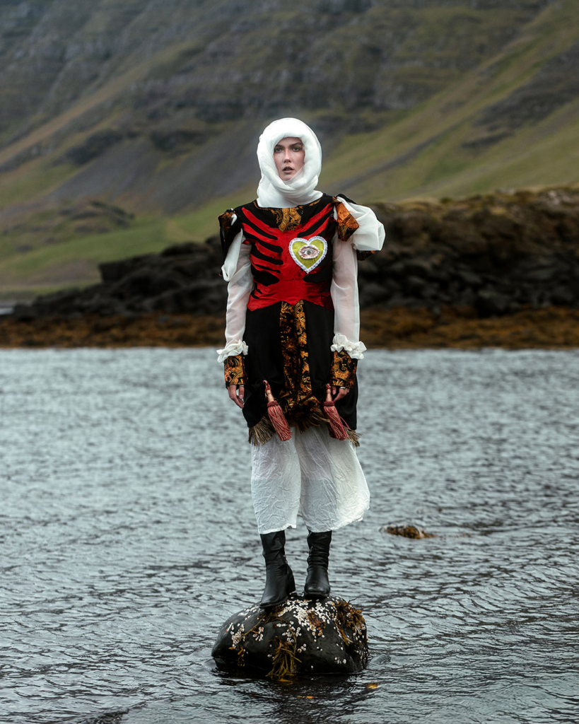 A woman stands on a rock in shallow water, wearing an elaborate black, red, and white costume with decorative embroidery and a white head covering. She faces the camera with a calm, steady expression, while the rugged Icelandic landscape and muted mountains behind her create a dramatic and cinematic atmosphere.