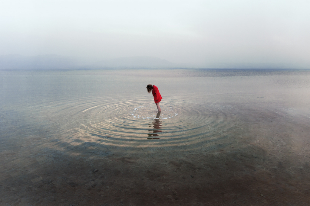 A woman in a bright red coat stands alone in shallow water, bending forward as concentric ripples spread outward around her feet. The vast, misty landscape and muted tones create a quiet, contemplative atmosphere, with the red garment standing out against the stillness of the scene.