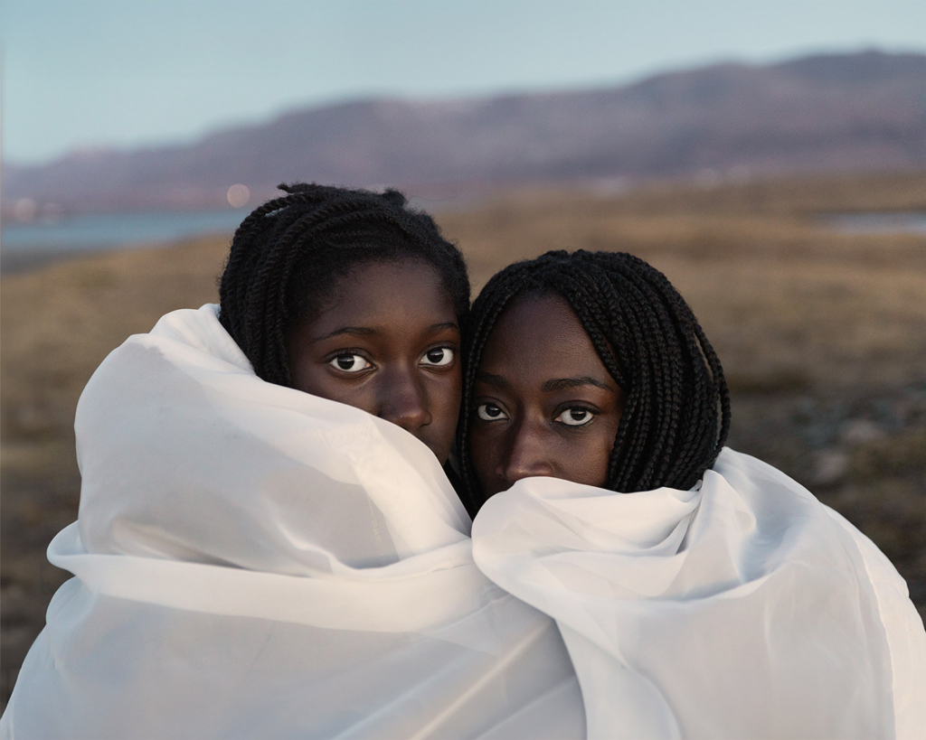 A mother and her son stand closely together outdoors, wrapped in flowing white fabric that frames their faces. They look directly at the camera with calm, thoughtful expressions, creating a sense of closeness and quiet connection against a softly blurred landscape of mountains and open land.