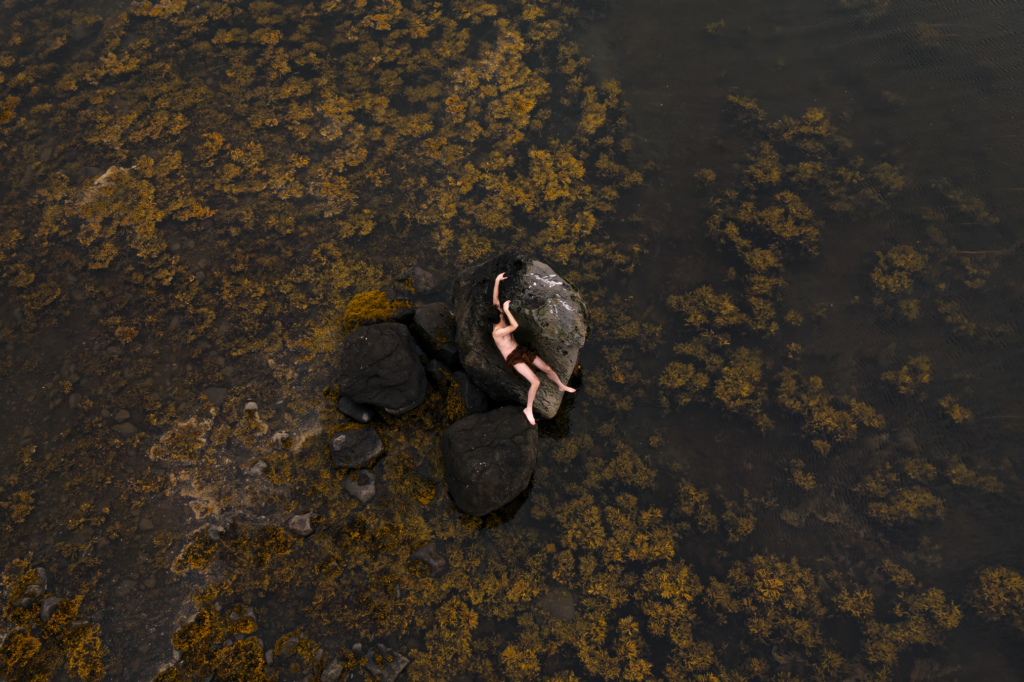 Aerial view of a woman lying across large coastal rocks surrounded by shallow water and golden-brown seaweed. Her body stretches across the stone in a relaxed pose, creating a striking contrast against the textured landscape and the calm, expansive shoreline.