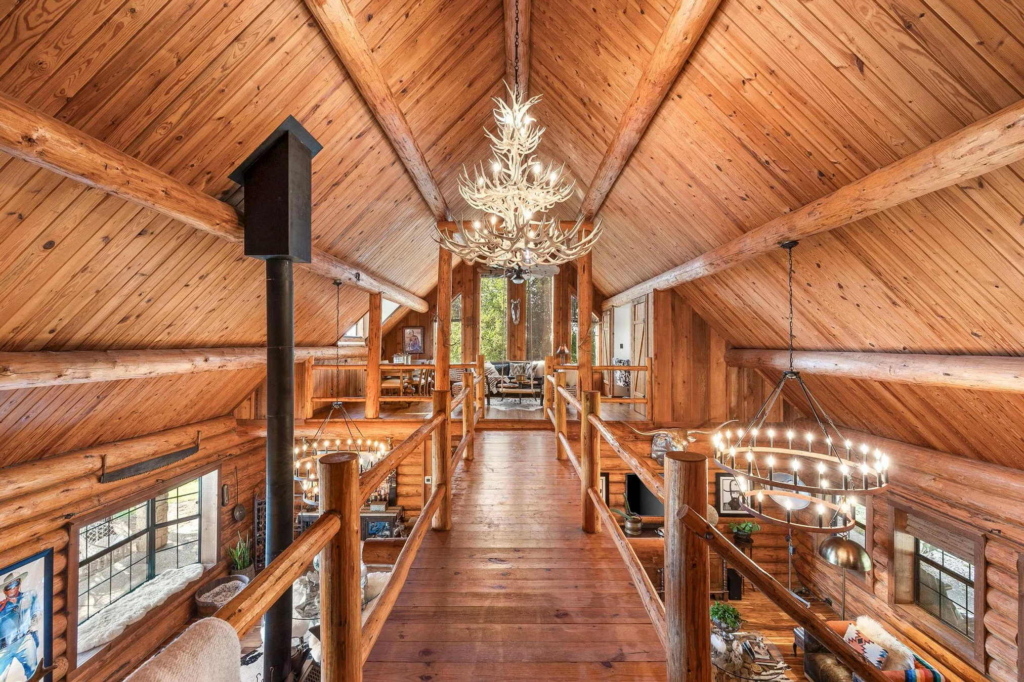 A spacious log cabin interior features exposed wooden beams, rustic chandeliers, and a loft walkway overlooking the living area below.
Warm natural wood tones and large windows create a cozy mountain-lodge atmosphere.